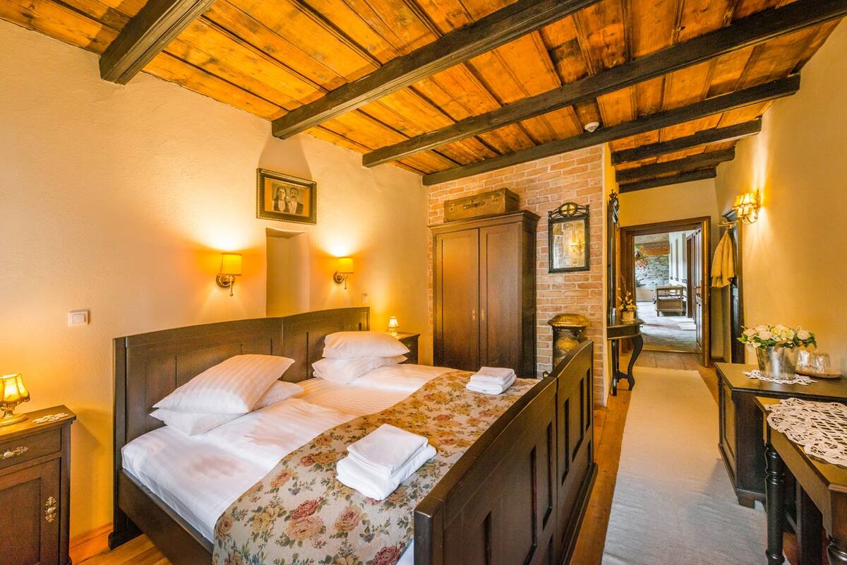 Habitación de hotel con techo de madera, una cama grande con ropa de cama floral, un armario de madera y una mesita con un jarrón de flores.