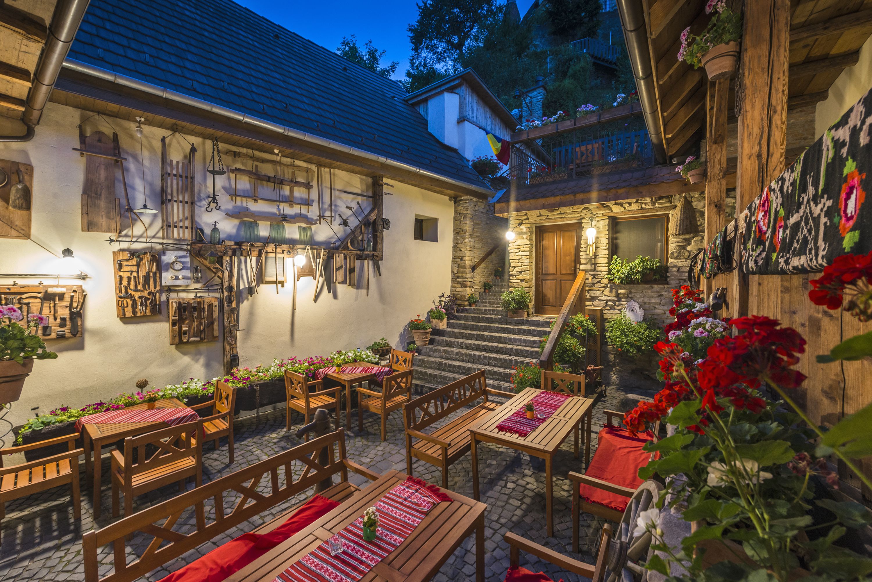 An outdoor patio area with wooden tables and chairs, stone steps leading to a wooden door, and various hanging items on the walls.