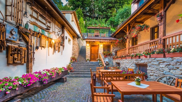 Un patio adoquinado con mesas y sillas de madera, paredes de piedra y jardineras con flores.