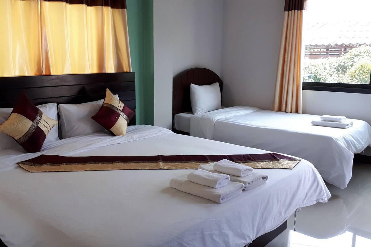 Habitación de hotel con dos camas, cada una con ropa blanca y almohadones decorativos. Una ventana grande con cortinas ofrece vistas a la vegetación exterior.