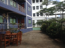 A courtyard with a wooden table and chairs, surrounded by potted plants and a blue building with balconies.