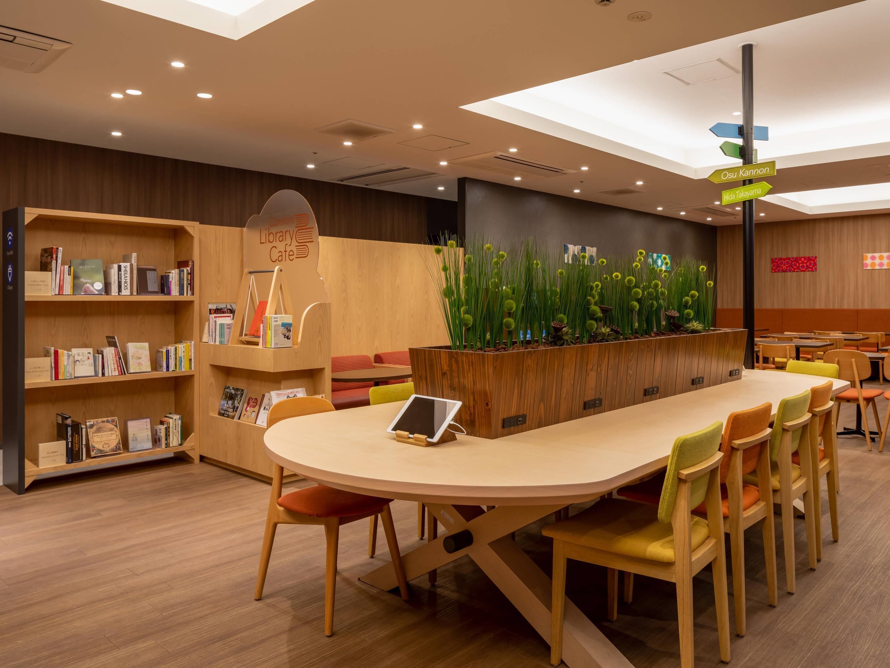 A modern library cafe with a long table, chairs, bookshelves, and a decorative plant wall.
