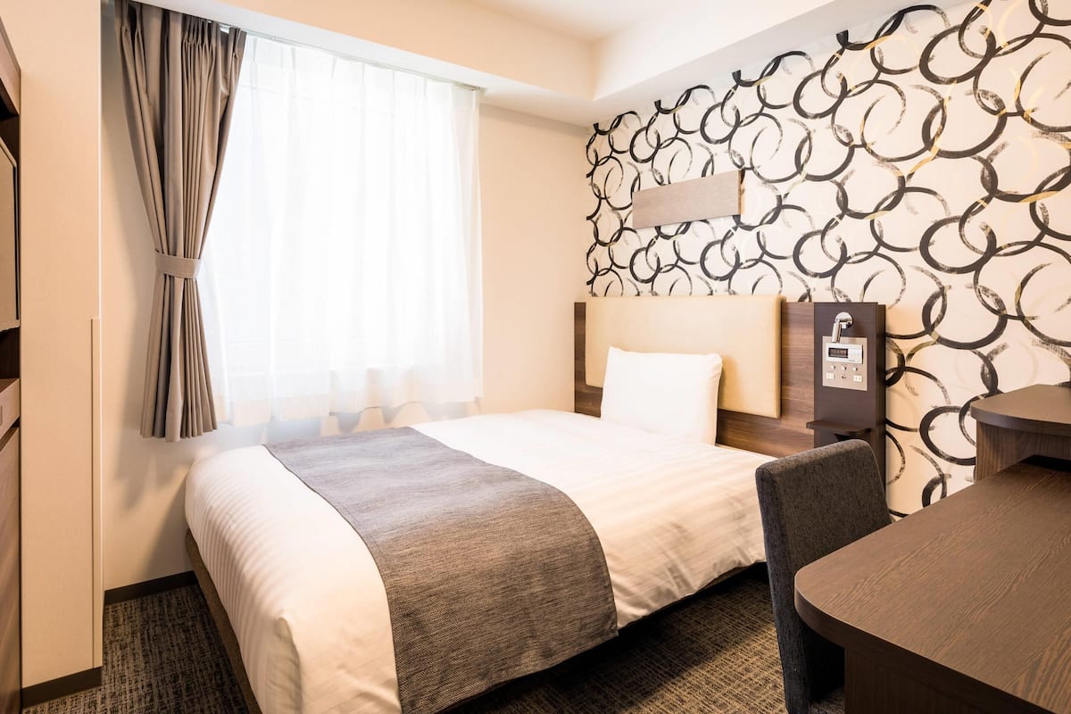 Une chambre d’hôtel comprenant un lit, un bureau, une chaise, une fenêtre avec des rideaux et un mur orné d’un motif circulaire.