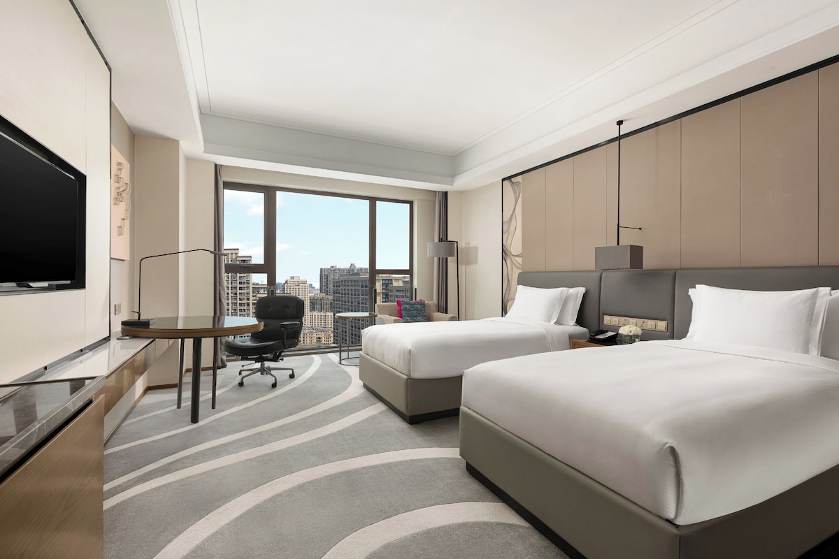 A hotel room with two beds, a desk, a chair, a TV, and a large window offering a city view.