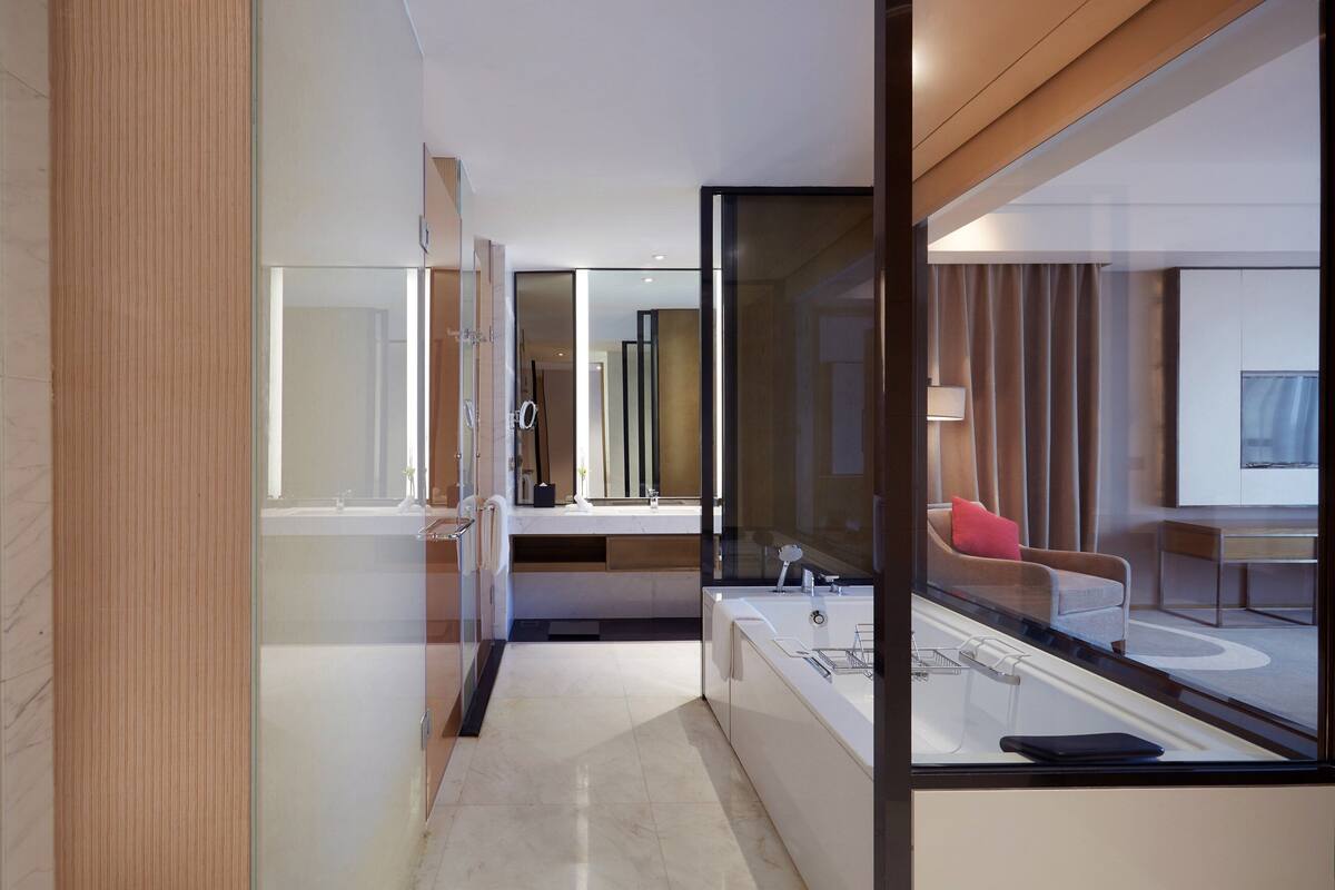 A modern hotel room with a glass partition, a bathtub, a sofa, and a dining area.