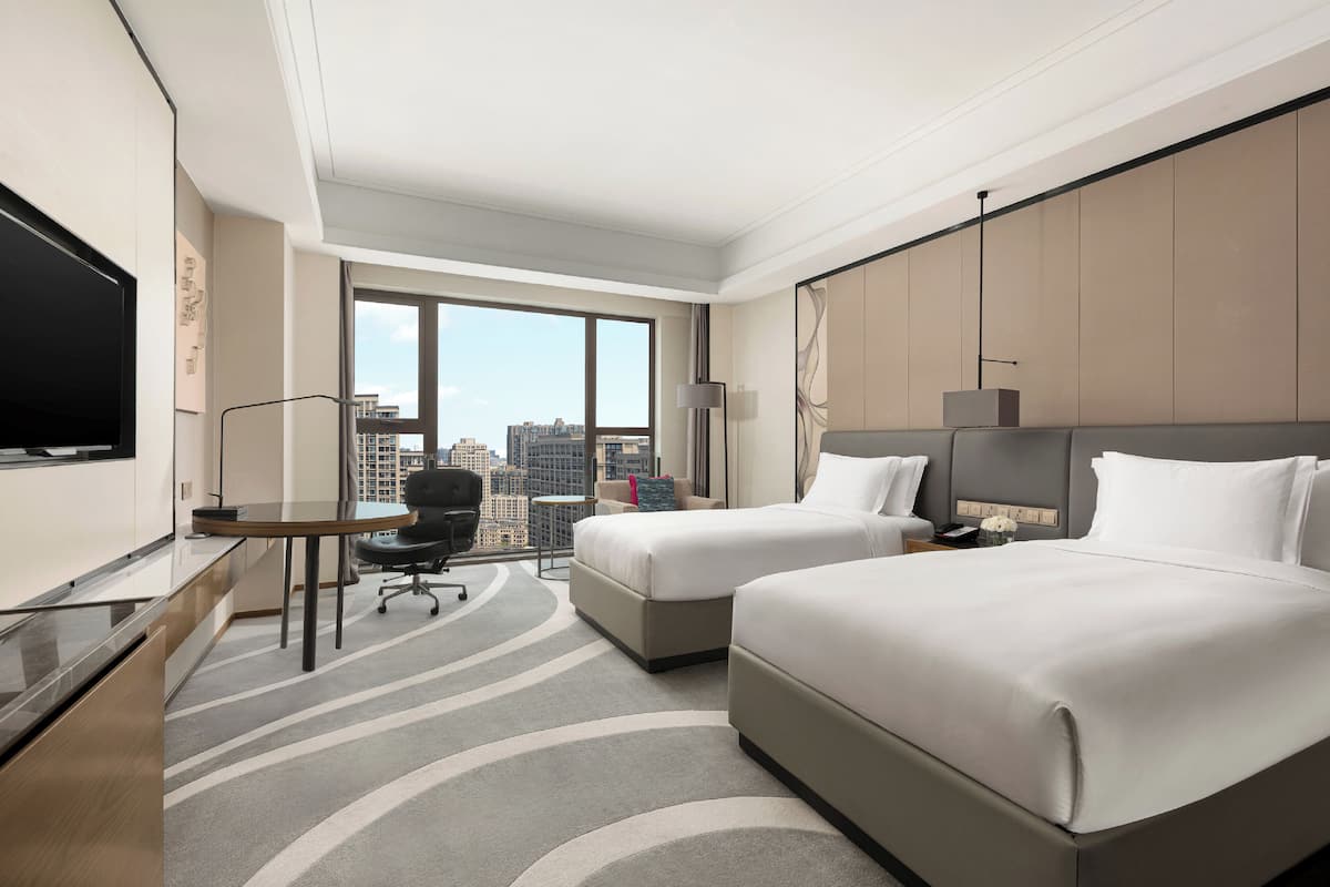 A hotel room with two beds, a desk, a chair, a TV, and a large window offering a city view.
