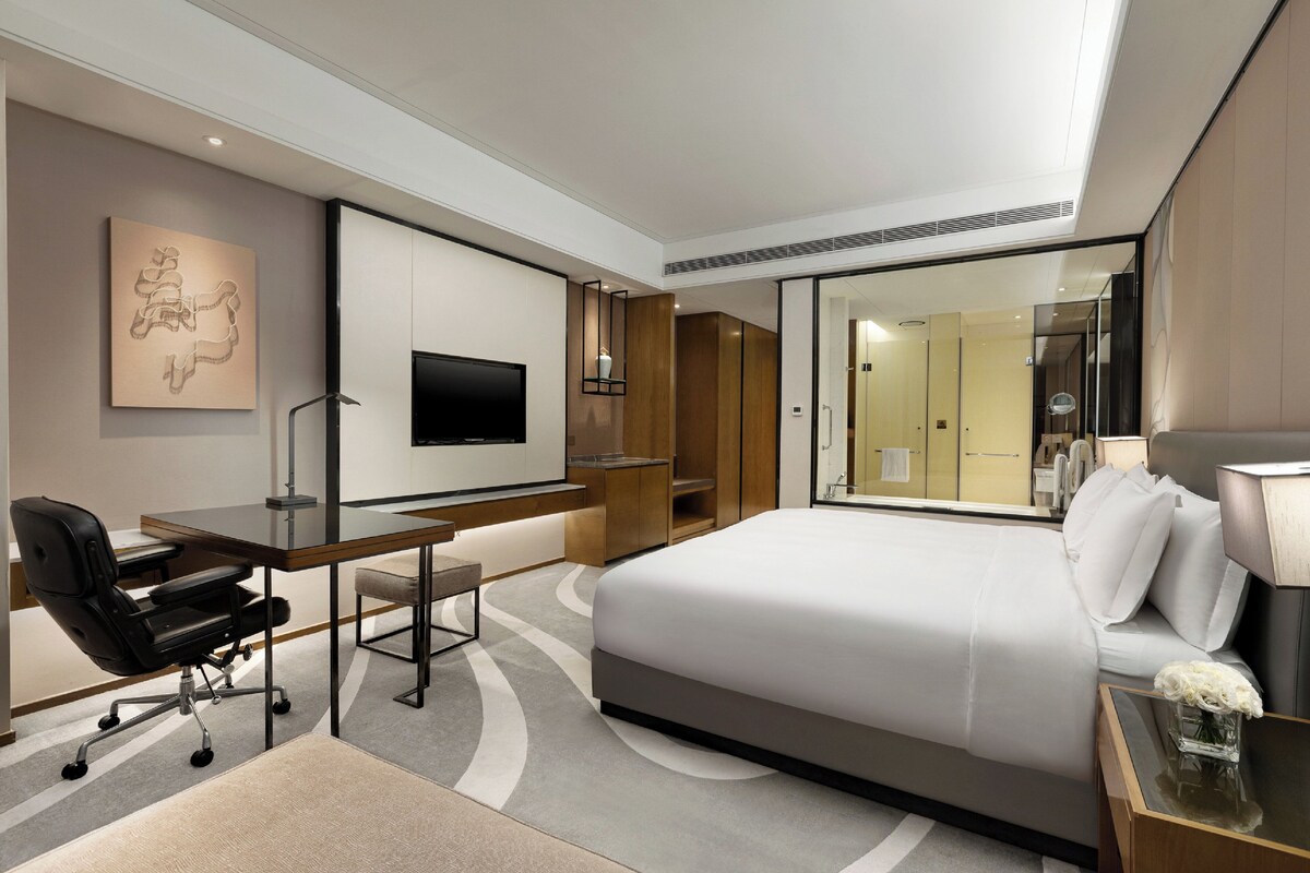 A modern hotel room with a large bed, a desk with a chair, a flat-screen TV, and a bathroom visible through a glass partition.