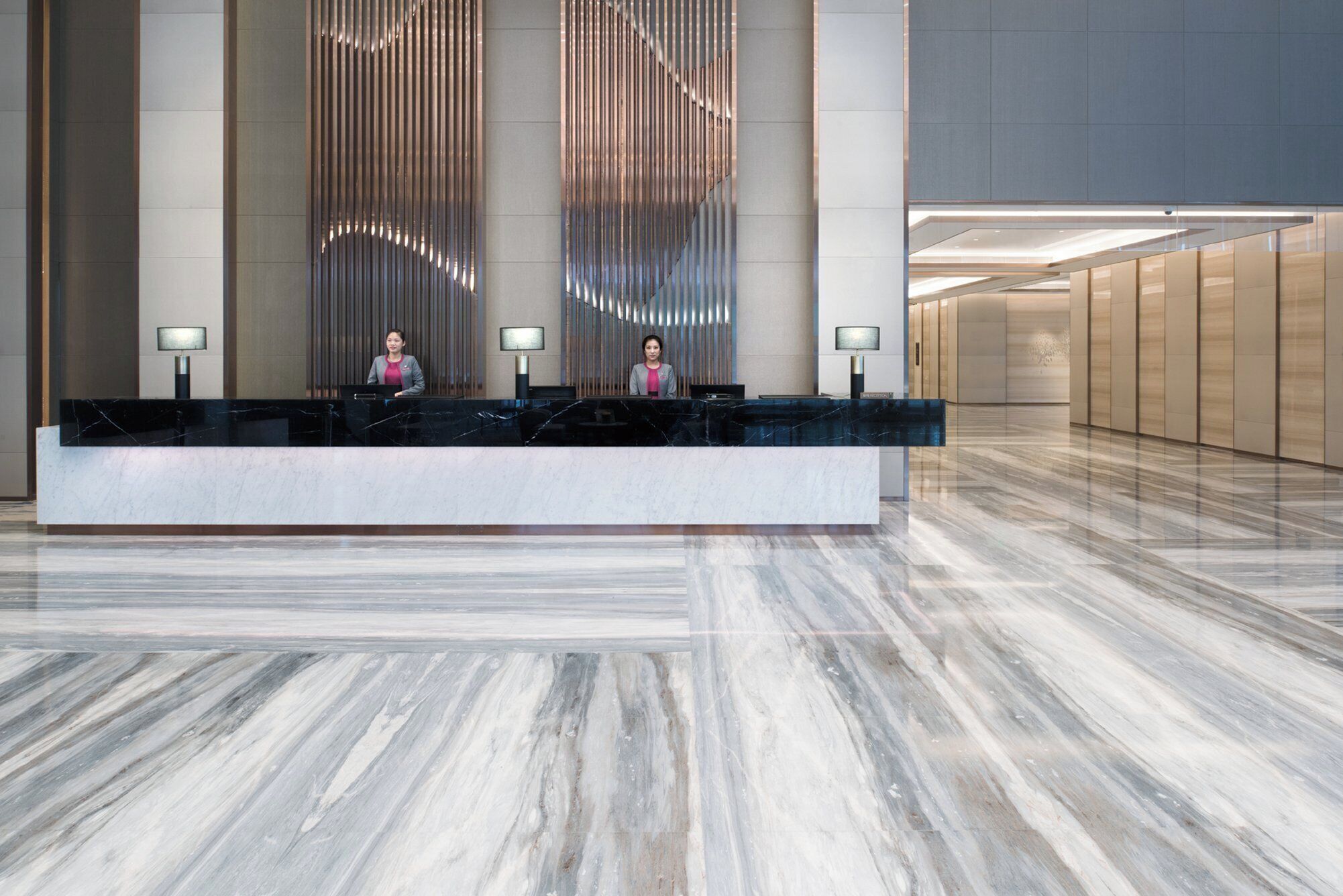 A hotel reception desk with two staff members, a modern interior design, and a marble floor.