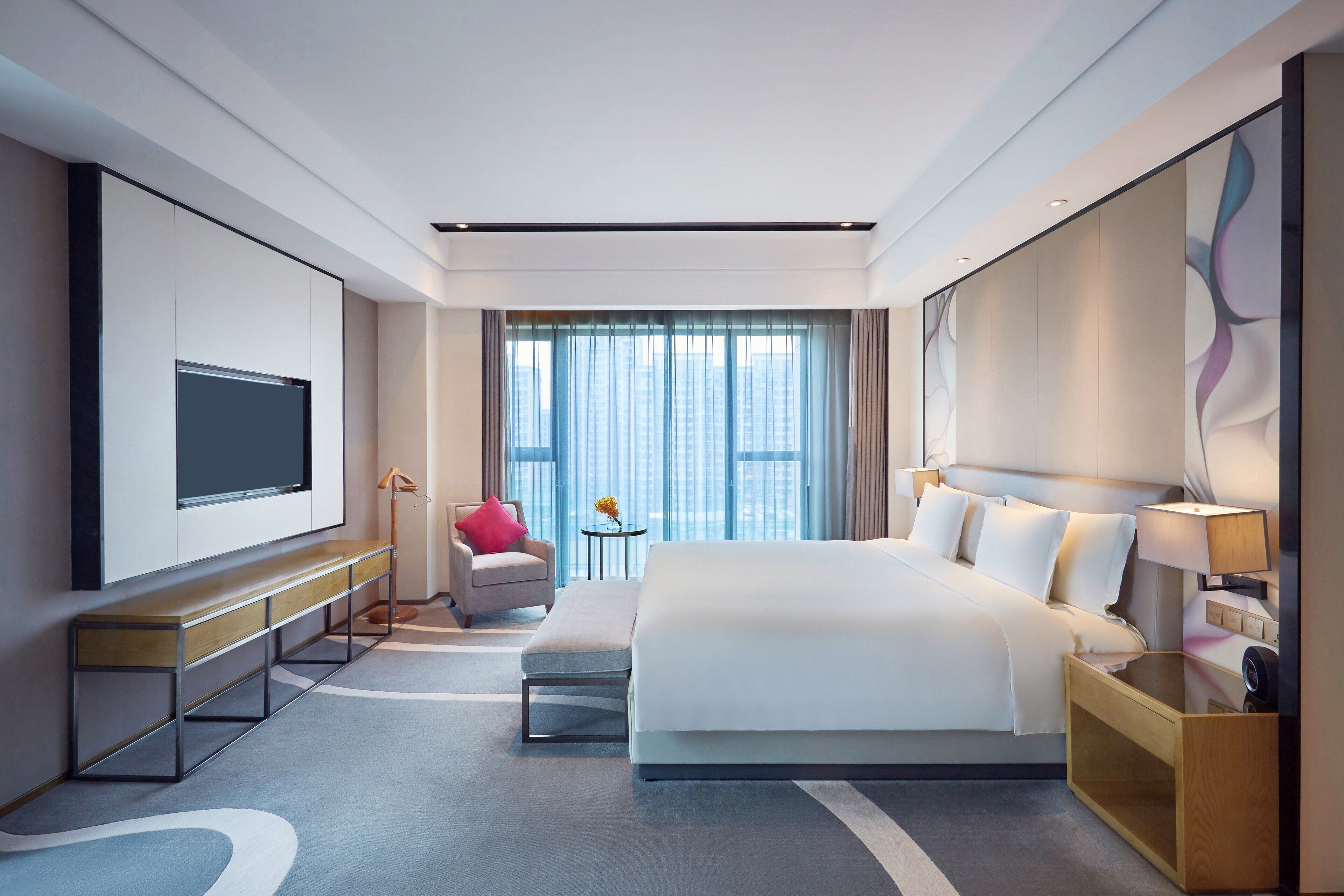 A modern hotel room with a large bed, a flat-screen TV, a bench, a small table with a lamp, and a view of the city through large windows.