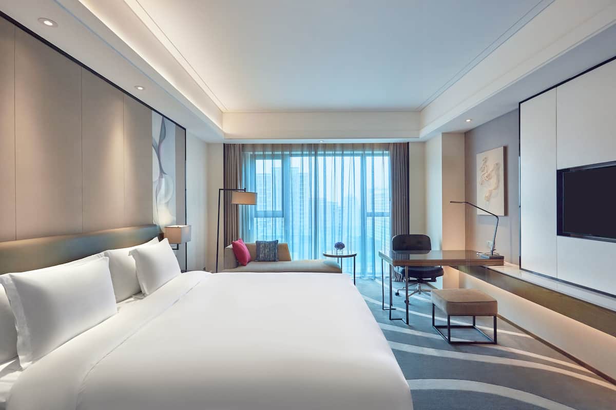 A modern hotel room with a large bed, a desk, a chair, and a TV on the wall.