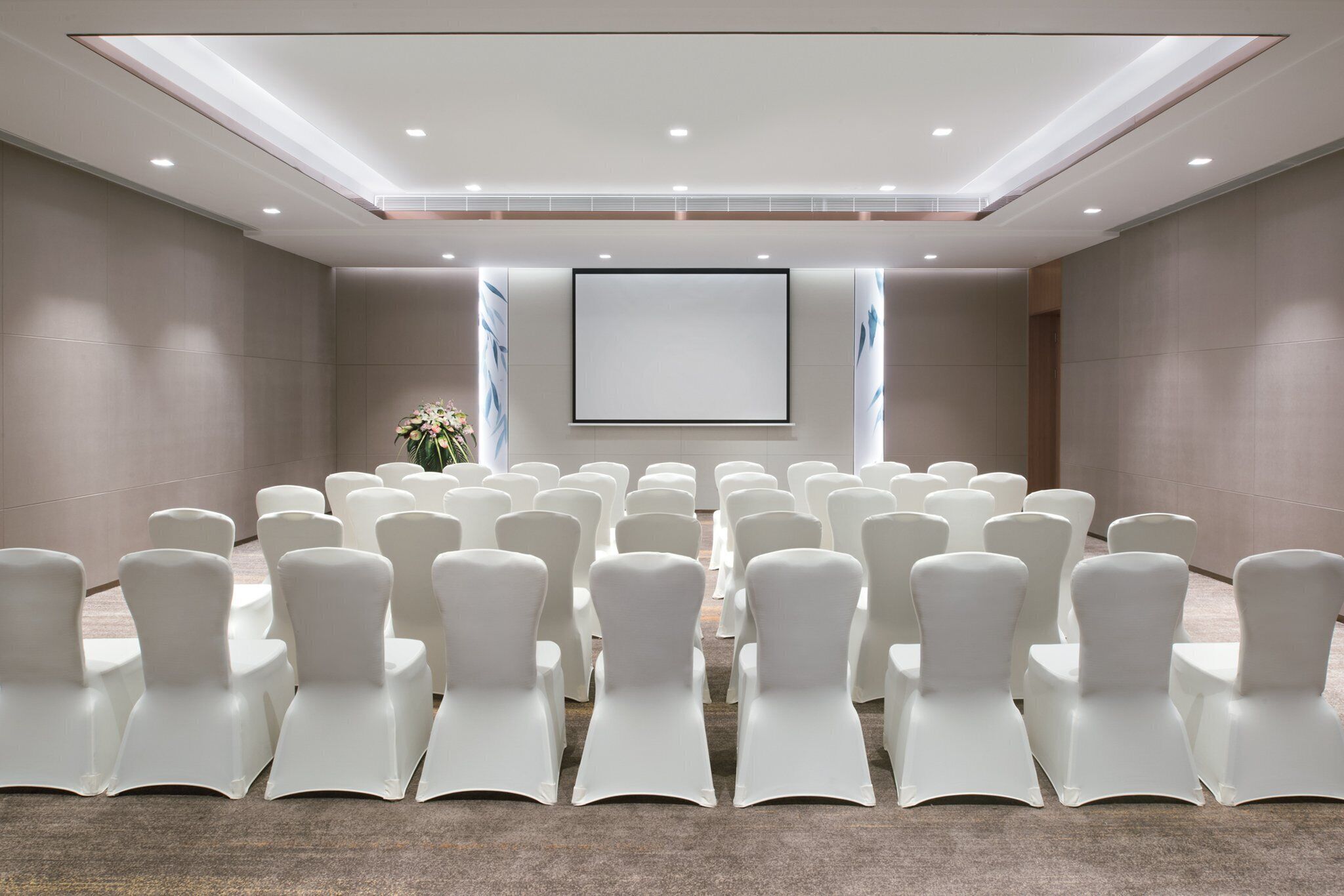 A conference room with rows of white chairs, a large screen, and a floral arrangement.