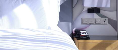 A hotel room with a bed, a bedside table with a lamp, a phone, and a power outlet.