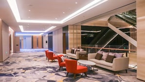 Eine moderne Hotelhalle mit Teppichboden, einer Sitzecke mit Sofas und Sesseln, einer Treppe und einem Glasgeländer.