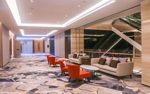 A modern hotel lobby with a carpeted floor, seating area with sofas and chairs, a staircase, and a glass railing.