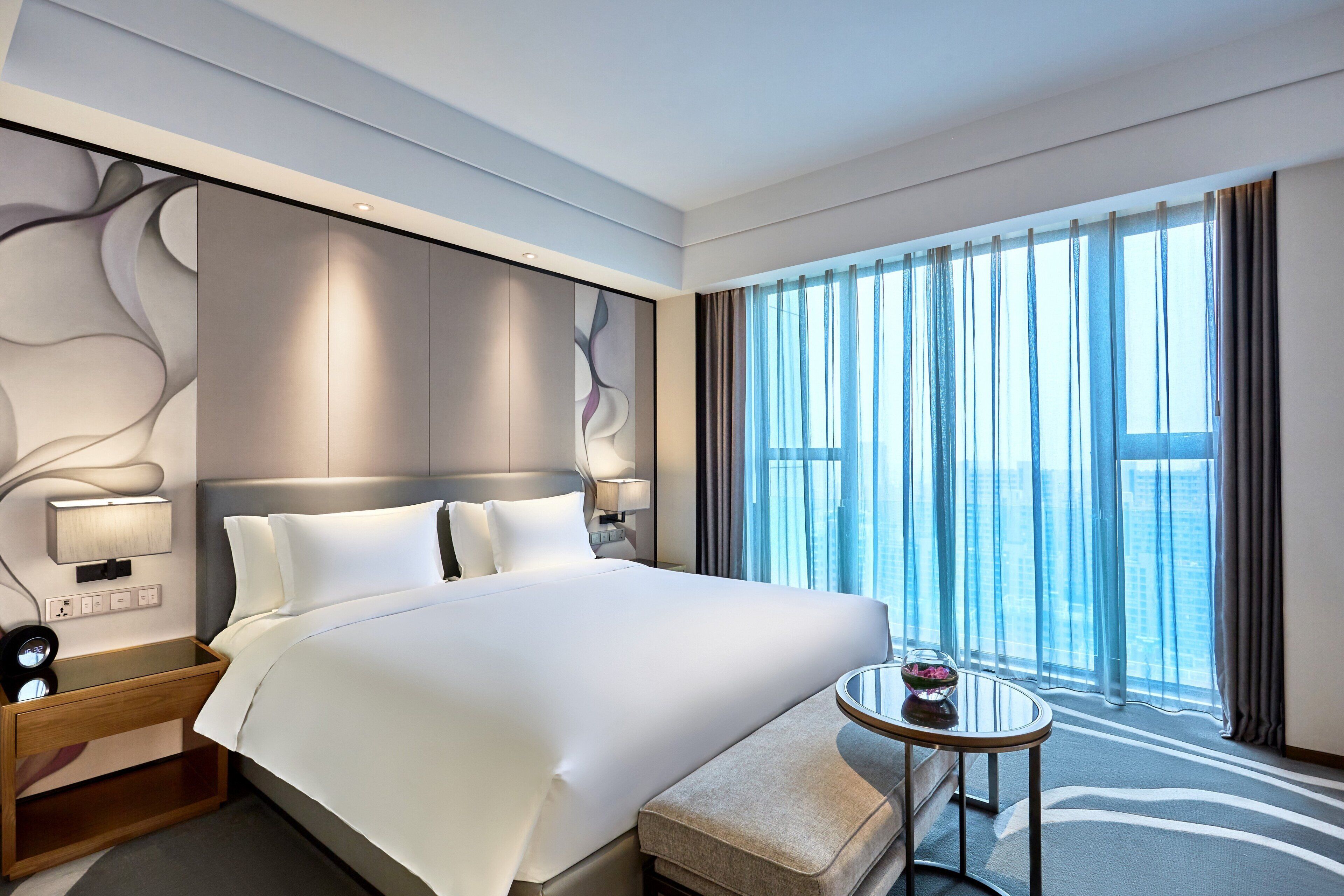 A modern hotel room with a large bed, bedside lamps, a small table, and a view of the city through large windows.
