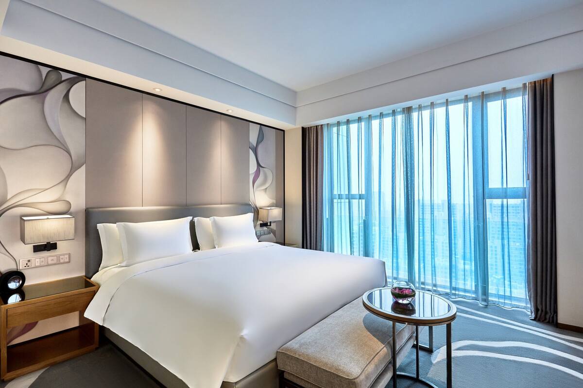 A modern hotel room with a large bed, a bedside table, a small round table, and a view of the city through large windows.