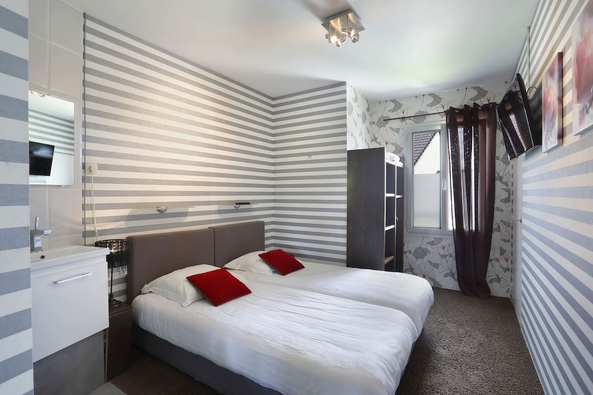 Una habitación de hotel con diseño de pared a rayas, una cama con ropa de cama blanca y almohadas rojas, una mesita de noche con lámpara y una ventana con cortinas.