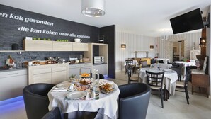 Un espace repas moderne avec une cuisine en arrière-plan, comprenant un tableau noir avec du texte, un lustre et un téléviseur à écran plat fixé au mur.