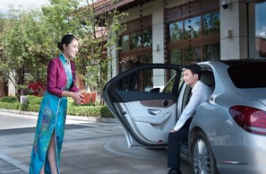 Eine Frau in traditioneller Kleidung steht neben einem Auto mit geöffneter Tür, während ein Mann im Inneren sitzt.