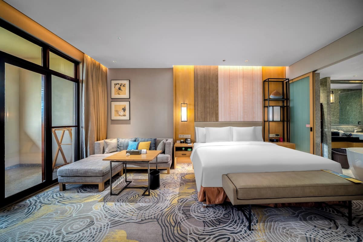 Une chambre d’hôtel avec un grand lit, un canapé, une petite table et un bureau. Les murs sont ornés de tableaux encadrés, il y a une lampe et une étagère avec des objets décoratifs.