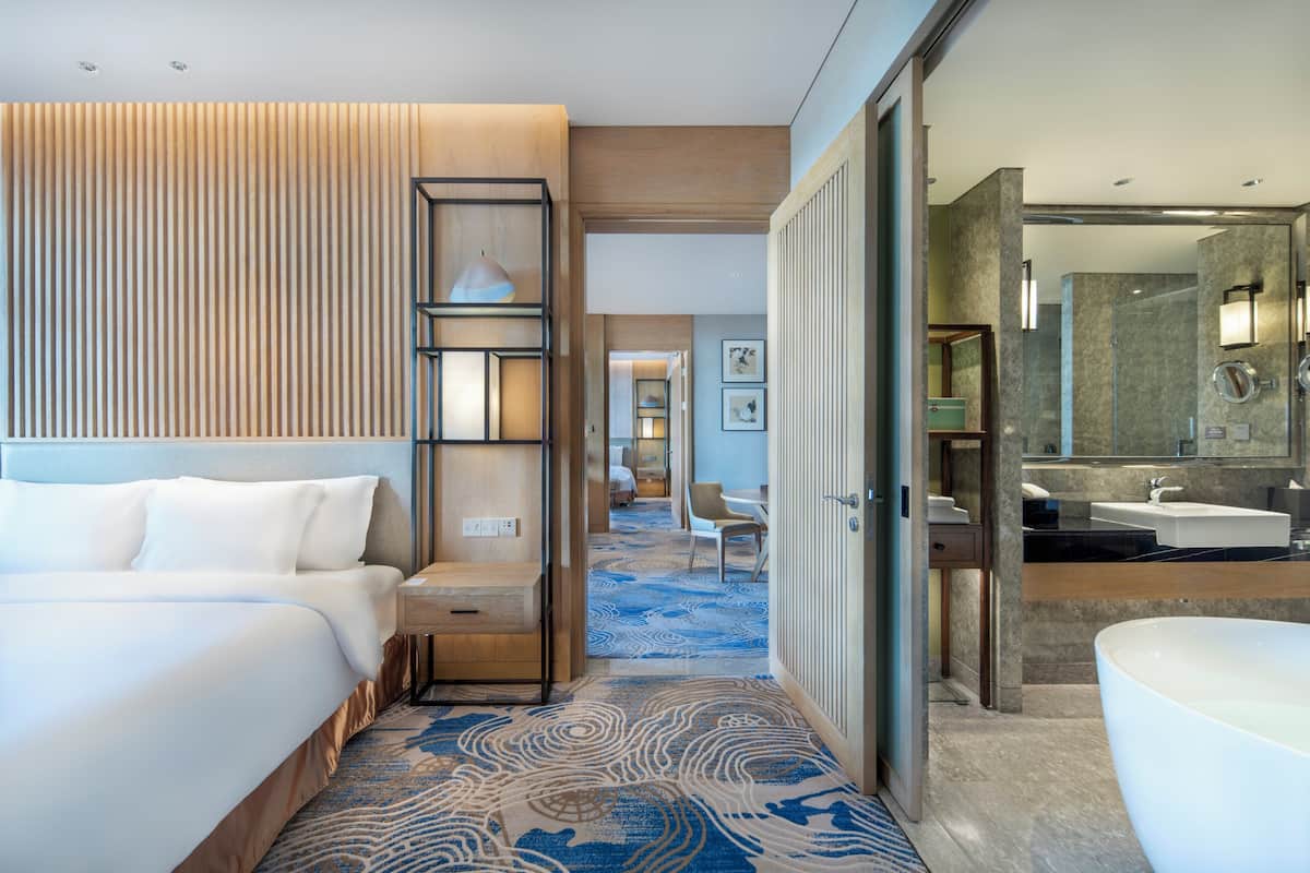 Une chambre d’hôtel avec un grand lit, un bureau, une chaise, une salle de bain avec une baignoire et un couloir menant à une autre chambre.