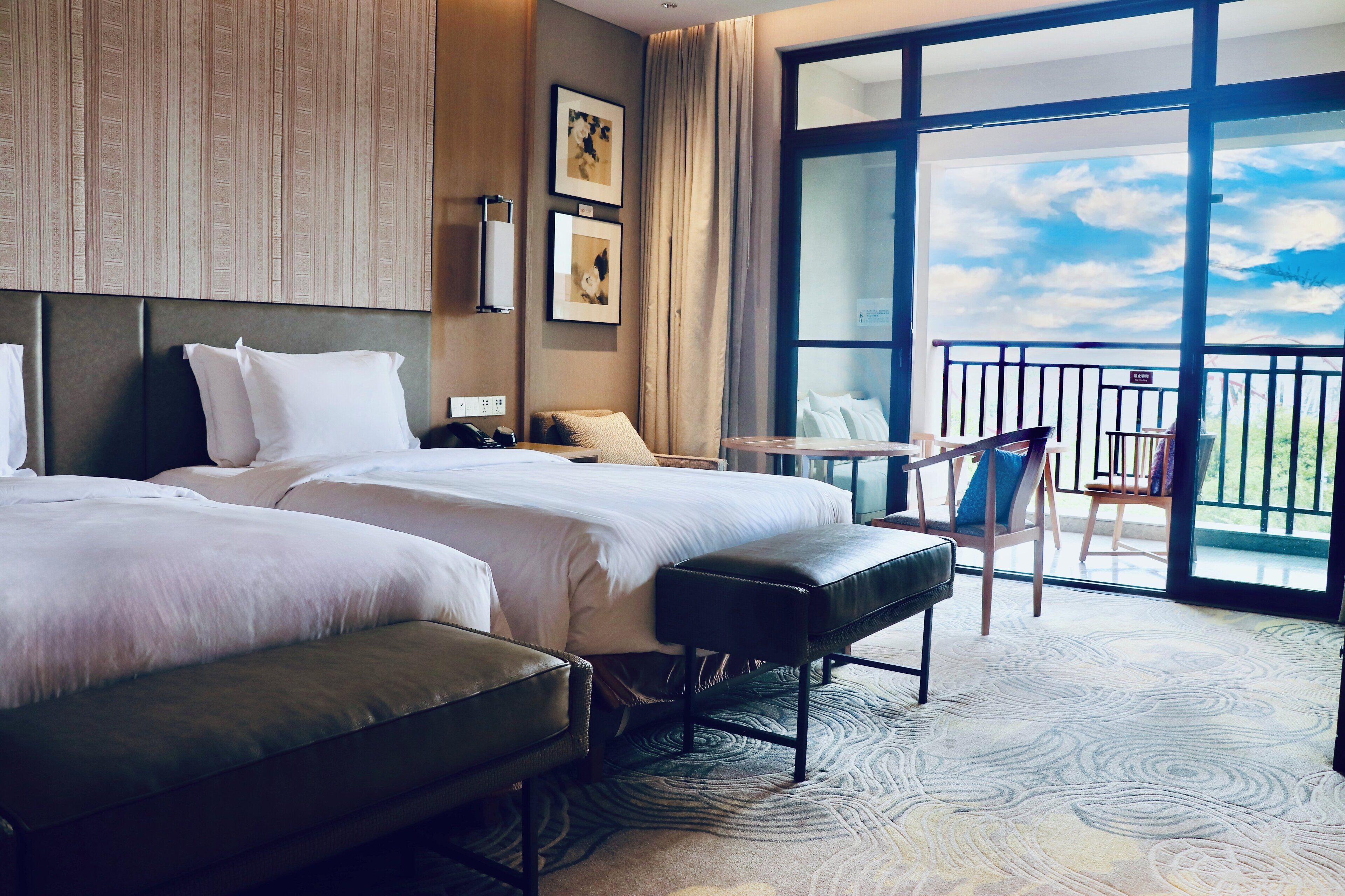 A hotel room with two beds, a bench, a small table, and a balcony with a view.