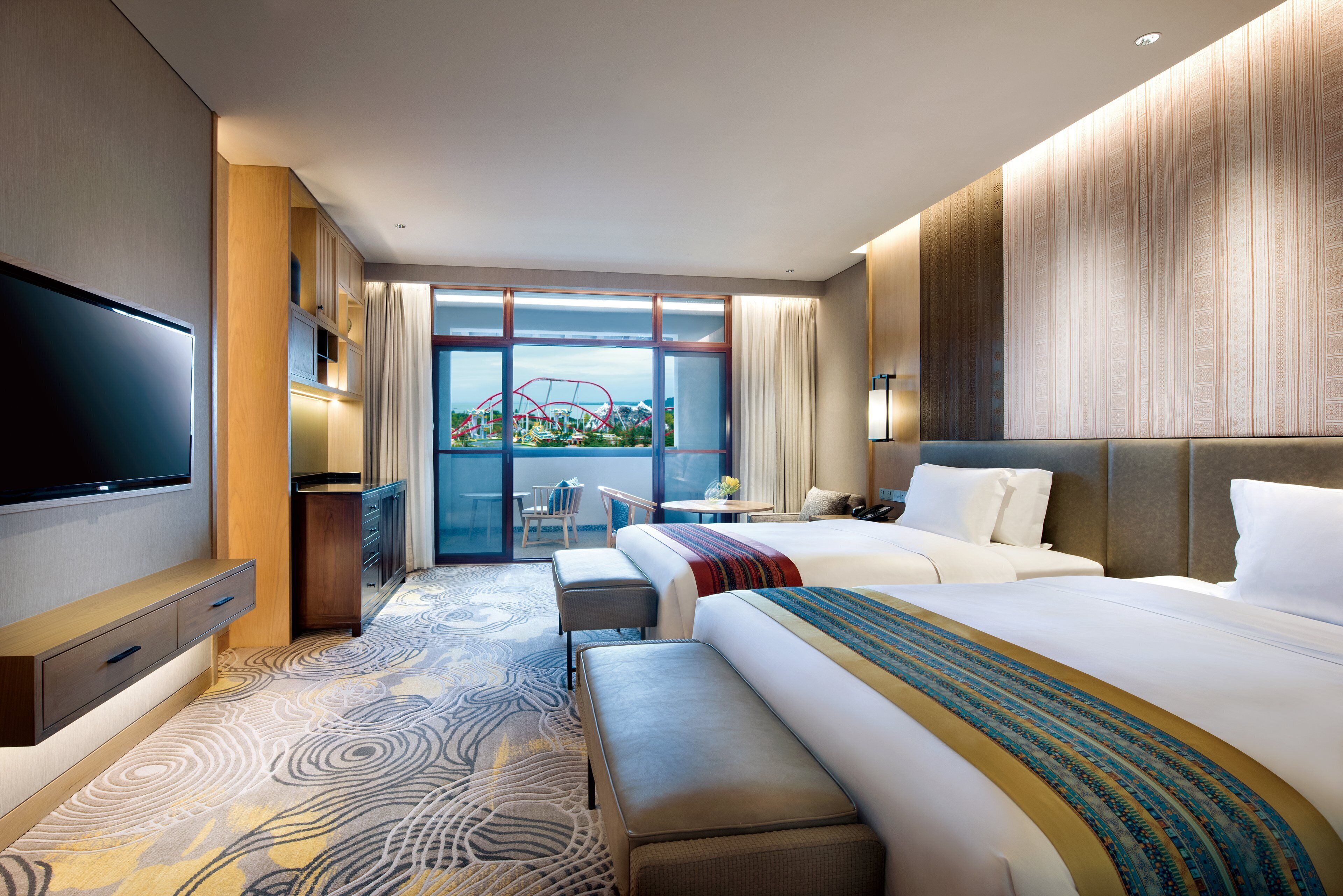 A modern hotel room with a large bed, a smaller bed, a flat-screen TV, a desk with a chair, and a balcony with a view of a cityscape.