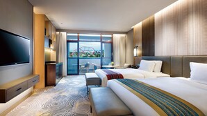 Une chambre d’hôtel moderne dotée d’un grand lit, d’un lit plus petit, d’un téléviseur à écran plat, d’un bureau avec une chaise et d’un balcon offrant une vue sur le paysage urbain.
