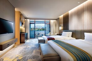 Une chambre d’hôtel moderne dotée d’un grand lit, d’un lit plus petit, d’un téléviseur à écran plat, d’un bureau avec une chaise et d’un balcon offrant une vue sur le paysage urbain.