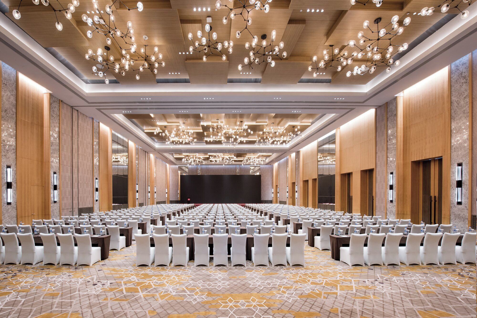 Un'ampia sala conferenze con file di tavoli e sedie, un grande schermo e lampadari moderni.