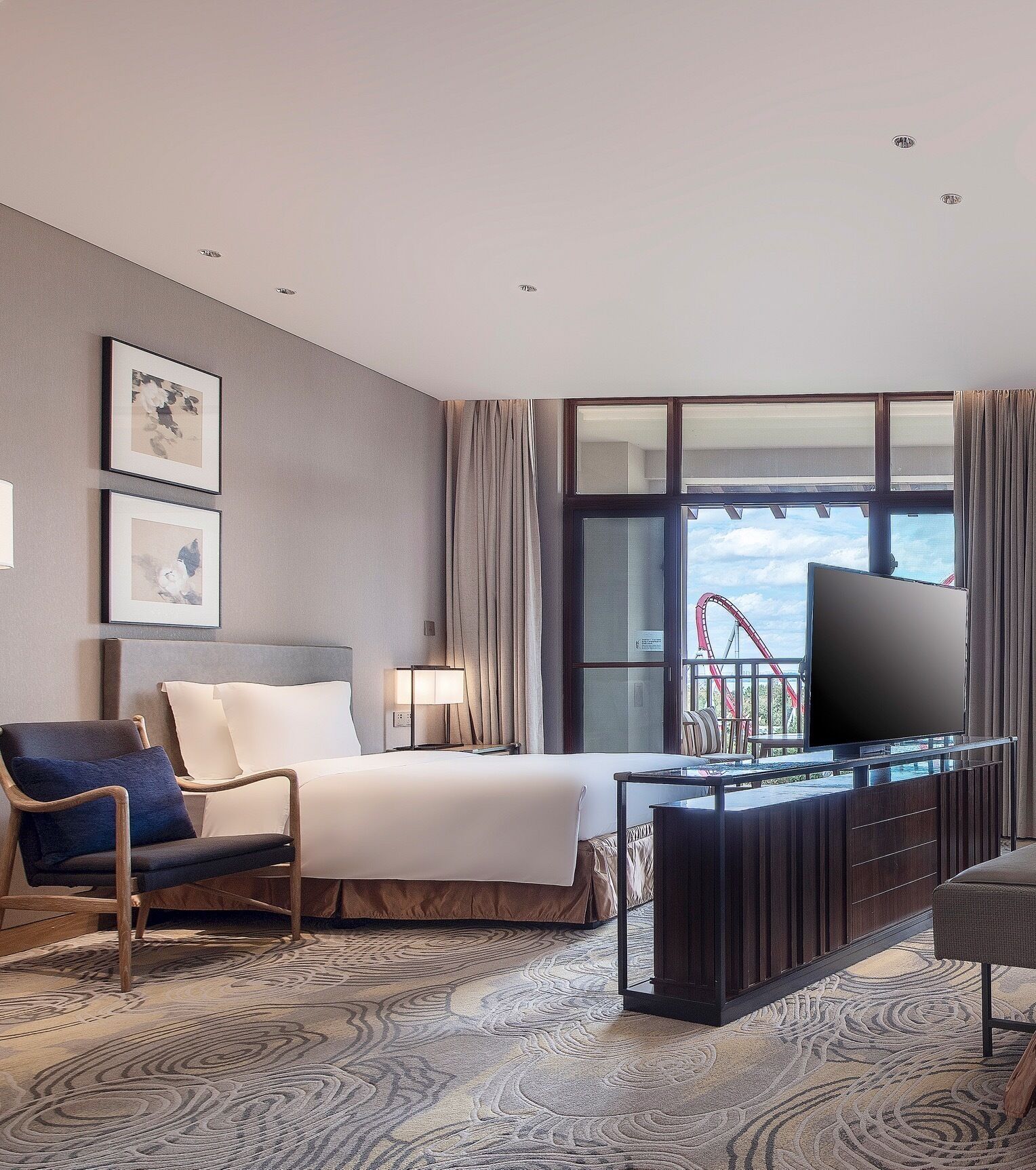 A hotel room with a bed, a chair, a TV, a desk, and a window with a view of a roller coaster.
