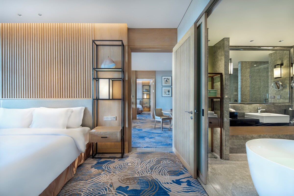 Ein Hotelzimmer mit einem großen Bett, einem Schreibtisch, einem Stuhl, einem Badezimmer mit Badewanne und einem Korridor, der in ein weiteres Zimmer führt.