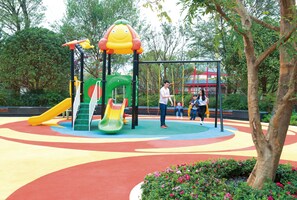 Ein Spielplatz mit Rutschen und Schaukeln, umgeben von Bäumen und einem roten Gebäude im Hintergrund.