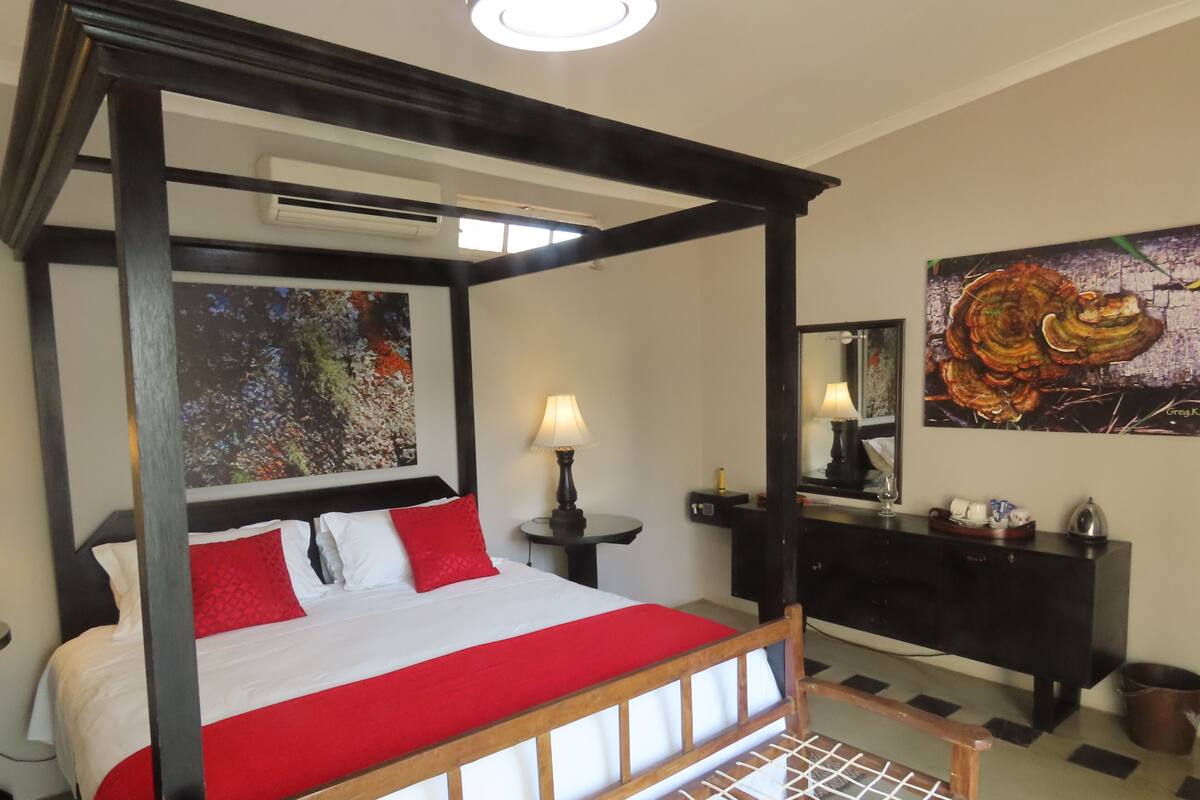 Un lit à baldaquin avec une couvre-lit rouge et blanche, une table de chevet avec une lampe, et un grand tableau abstrait accroché au mur.