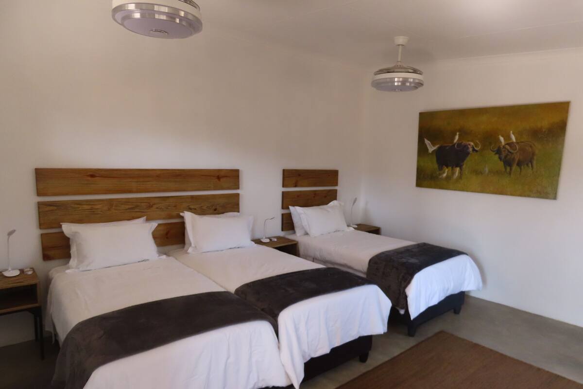 Une chambre d’hôtel avec deux lits, des têtes de lit en bois, du linge de lit blanc, un tableau représentant des taureaux et un ventilateur de plafond.