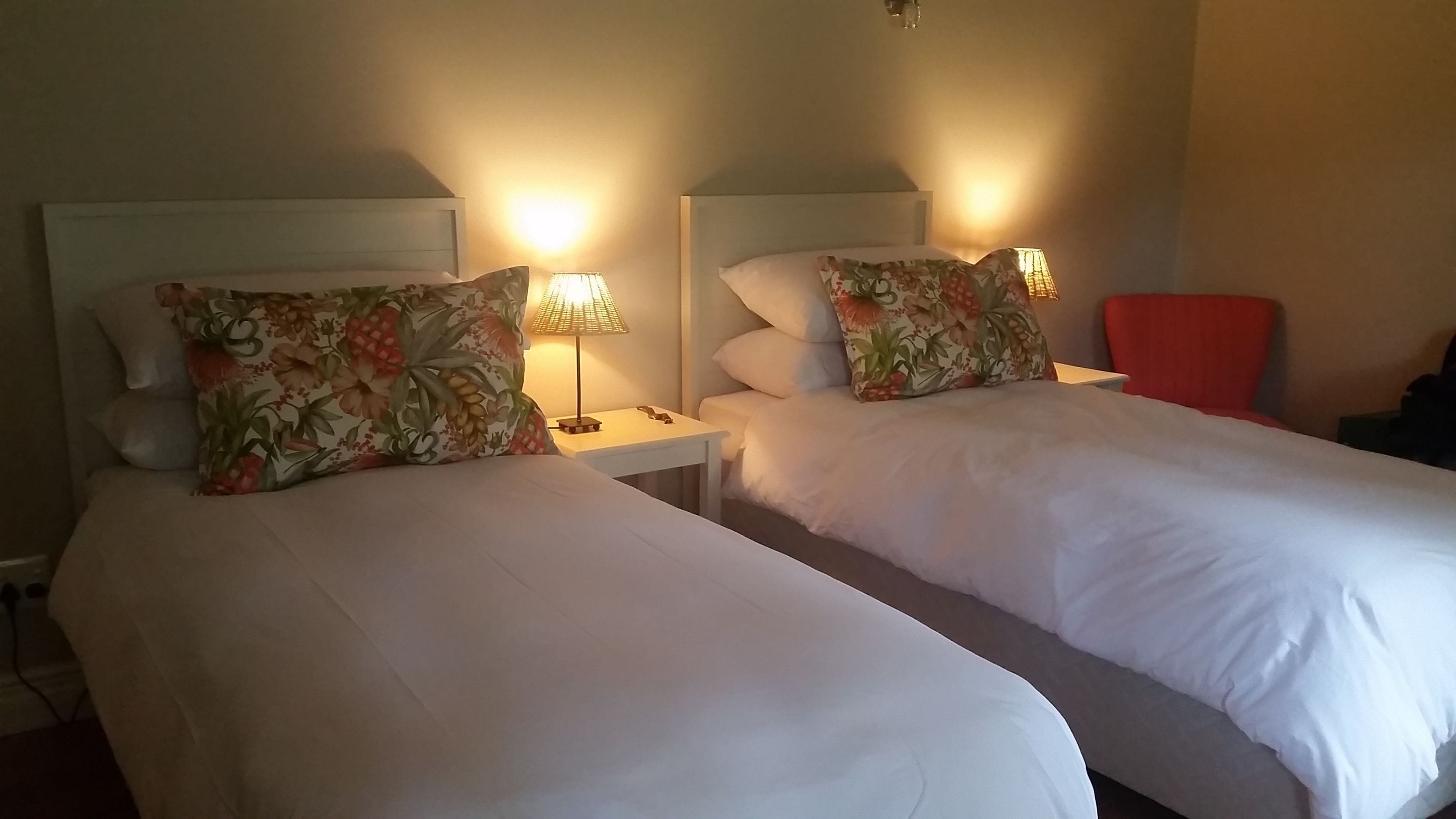 A hotel room with two beds, each with floral-patterned pillows and white bedding. There is a nightstand with a lamp between the beds, and a red chair in the corner.