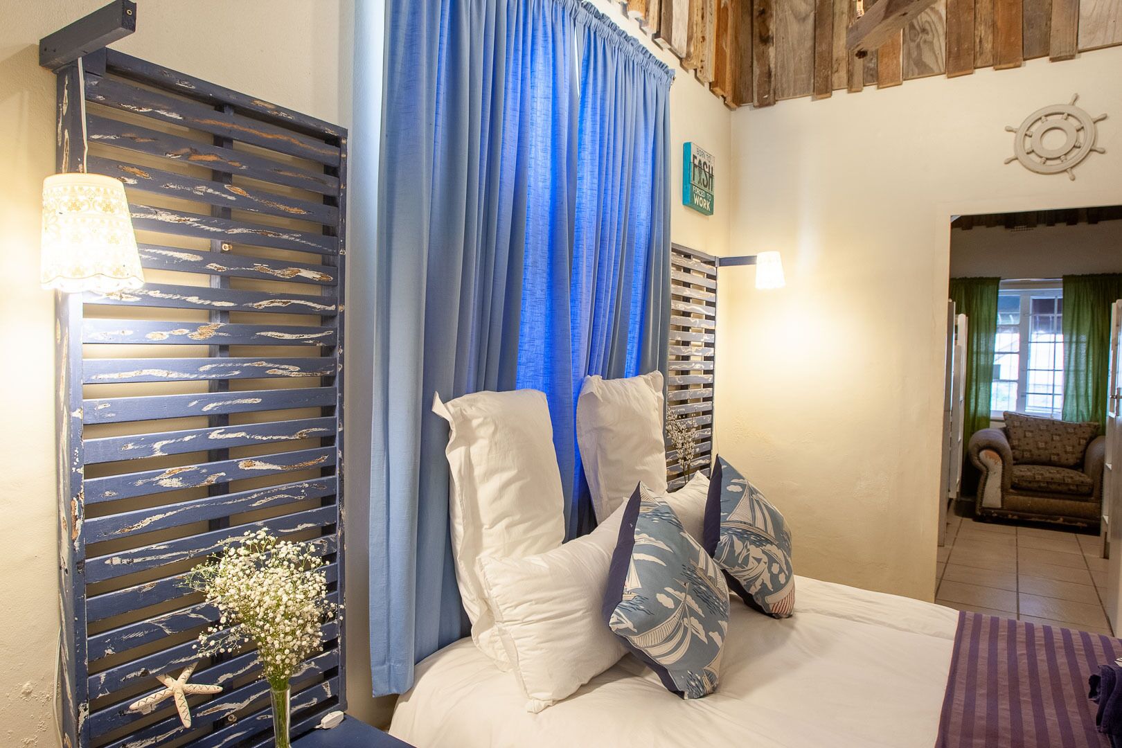 Een slaapkamer met een bed, een blauw gordijn, een houten hoofdeinde, een nachtkastje met een lamp en een aan de muur bevestigd scheepswiel.