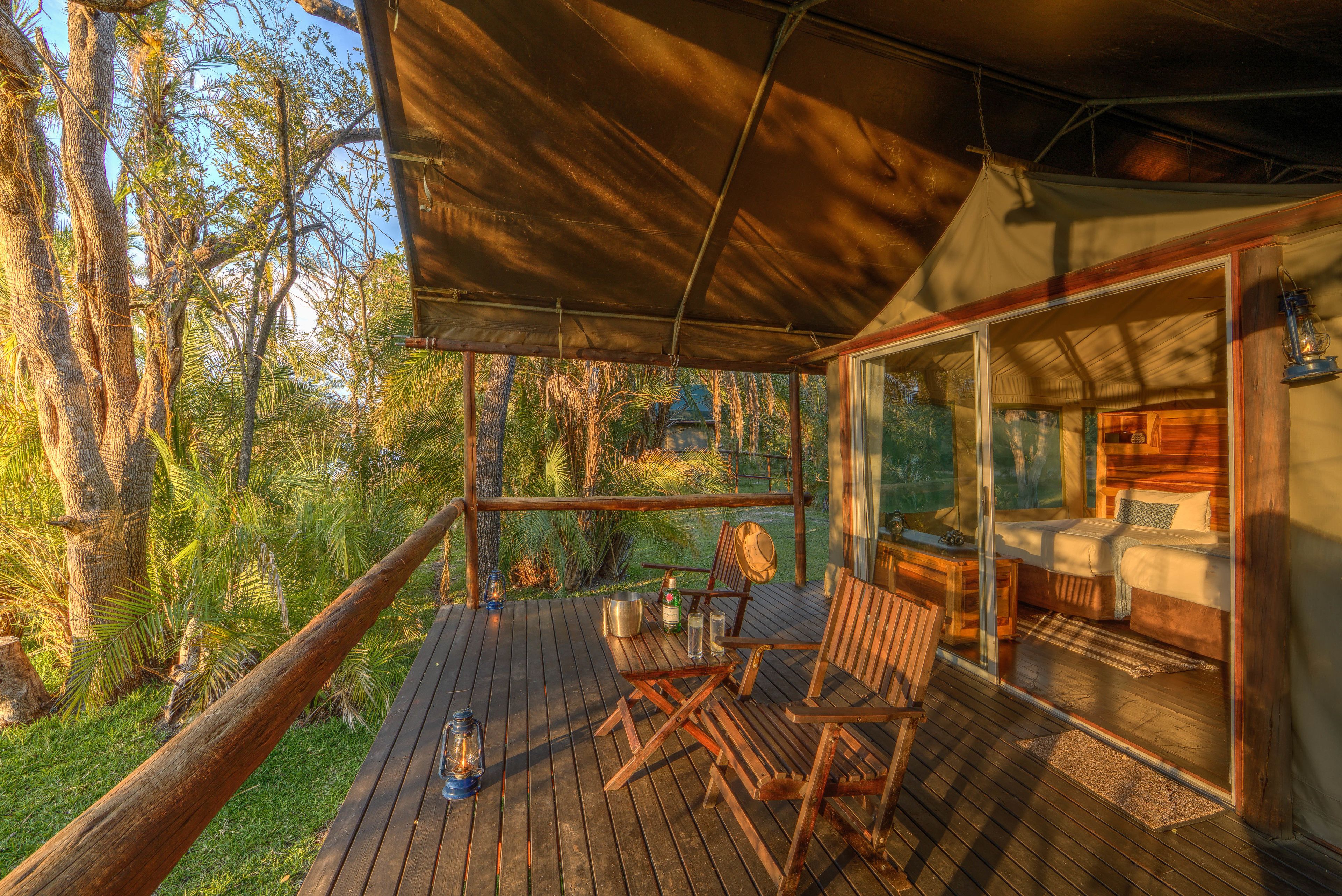 A wooden deck with a glass-enclosed bedroom, a dining area with chairs and a table, and a view of the surrounding forest.