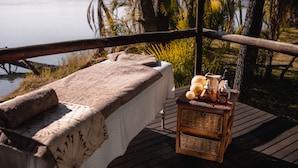 Een houten terras met een massagetafel bedekt met een witte handdoek, een klein houten bijzettafeltje met flessen en een kaars, en uitzicht op een meer en bomen.