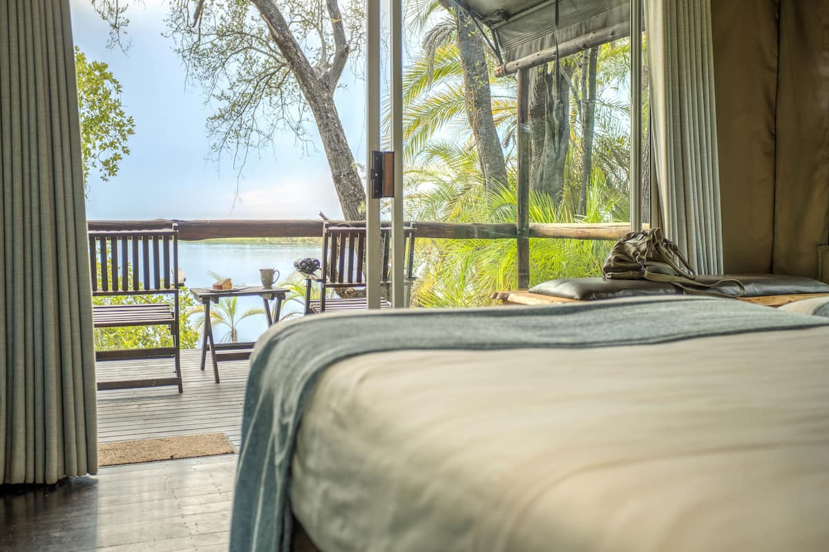 A bedroom with a view of a lake, a wooden deck, and a table with a chair.