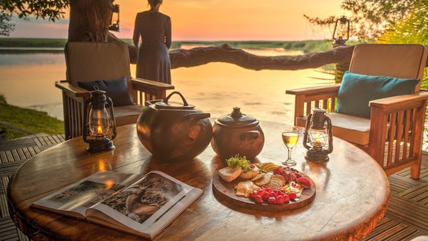 Een houten tafel met een boek, theepot en lekkernijen, met uitzicht op een wateroppervlak tijdens de zonsondergang.