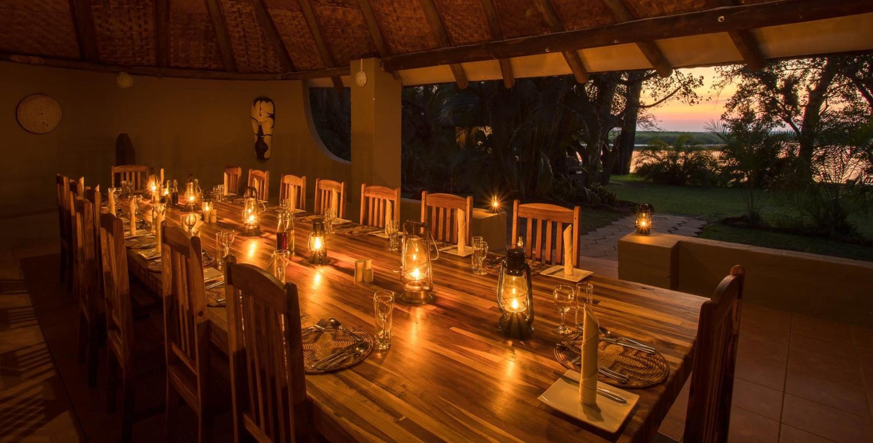 A long wooden dining table set for a meal with candles and lanterns, surrounded by wooden chairs, under a thatched roof.