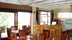 Un espace repas avec des tables et des chaises en bois, un coin bar avec un comptoir et des tabourets, et de grandes fenêtres laissant entrer la lumière naturelle.