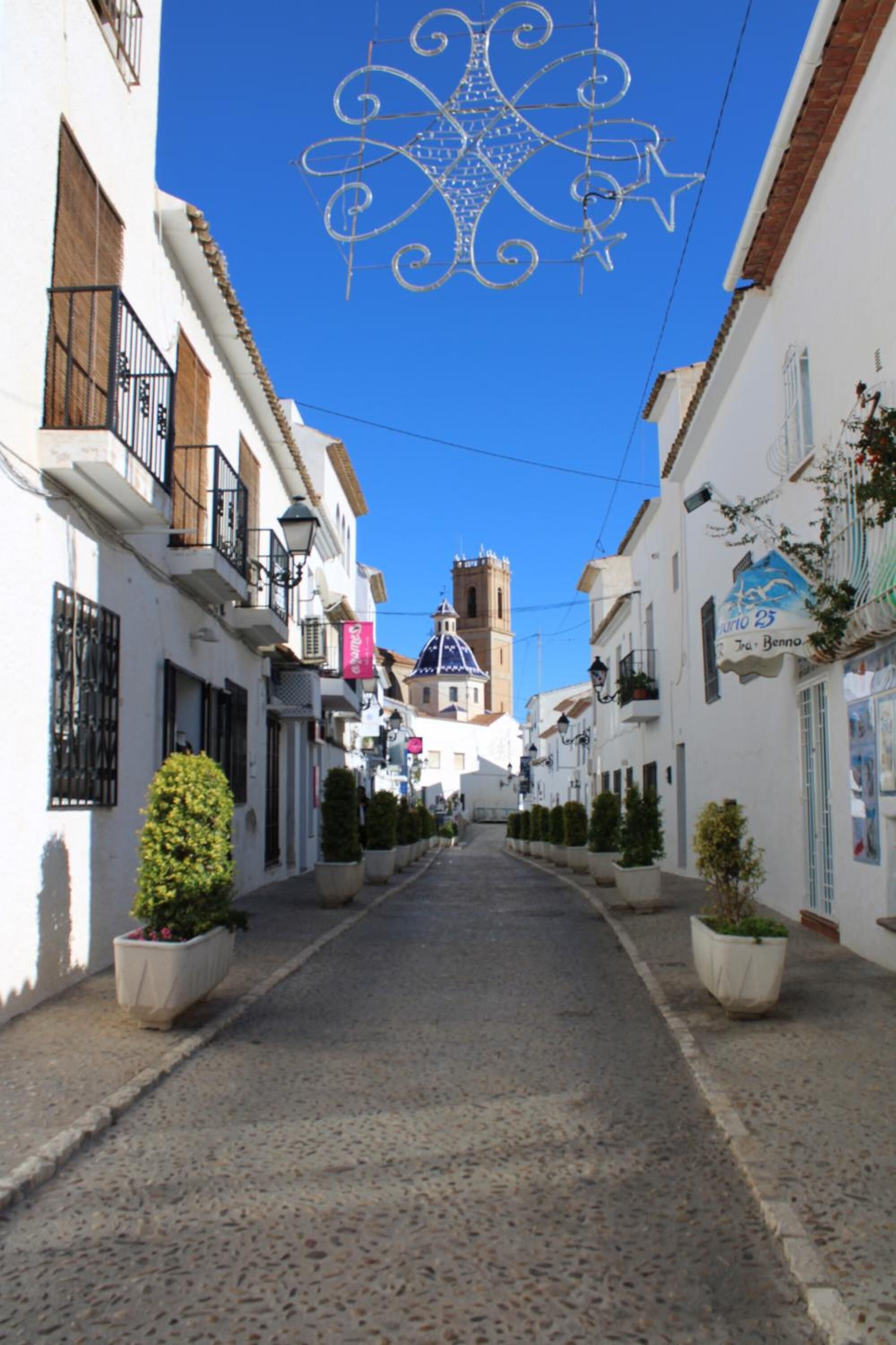 Una calle estrecha flanqueada por edificios blancos, algunos con balcones y plantas. Una estructura metálica decorativa cuelga sobre la calle.