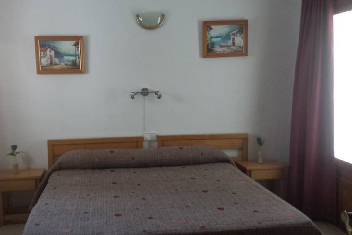 A bedroom with a bed, two bedside tables, a nightstand, a wall-mounted lamp, and two framed pictures.