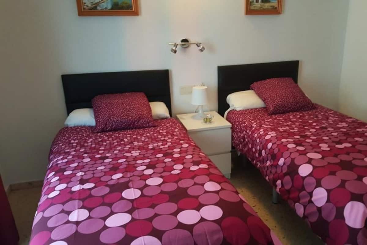 A room with two beds, each with a red and white polka dot bedspread, white nightstands, and a wall-mounted lamp.