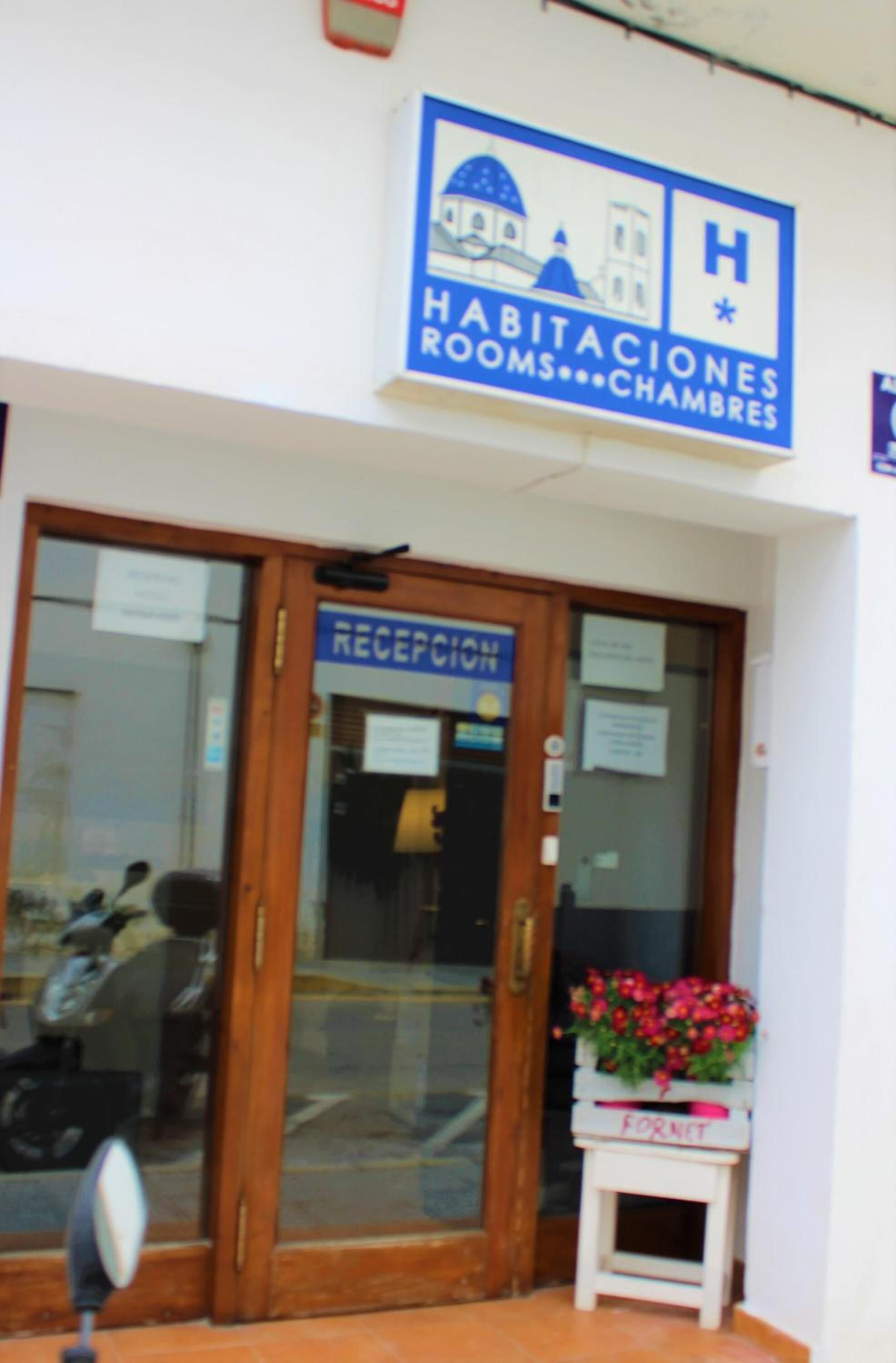 A hotel entrance with a reception desk, a sign indicating 'HABITACIONES ROOMS...CHAMBERS', and a small planter with flowers.