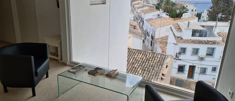 Un salon moderne avec une table basse en verre, deux fauteuils noirs et une vue sur une ville côtière.