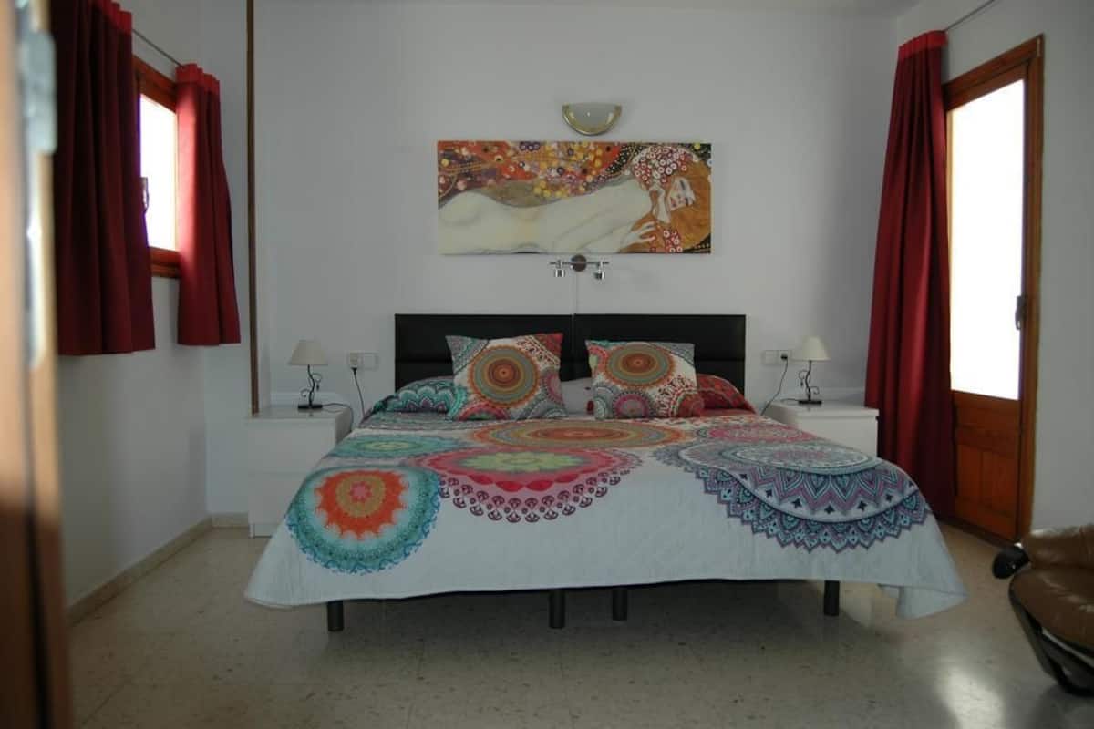 A bedroom with a bed, two bedside tables, a nightstand, a wall-mounted lamp, and a painting.