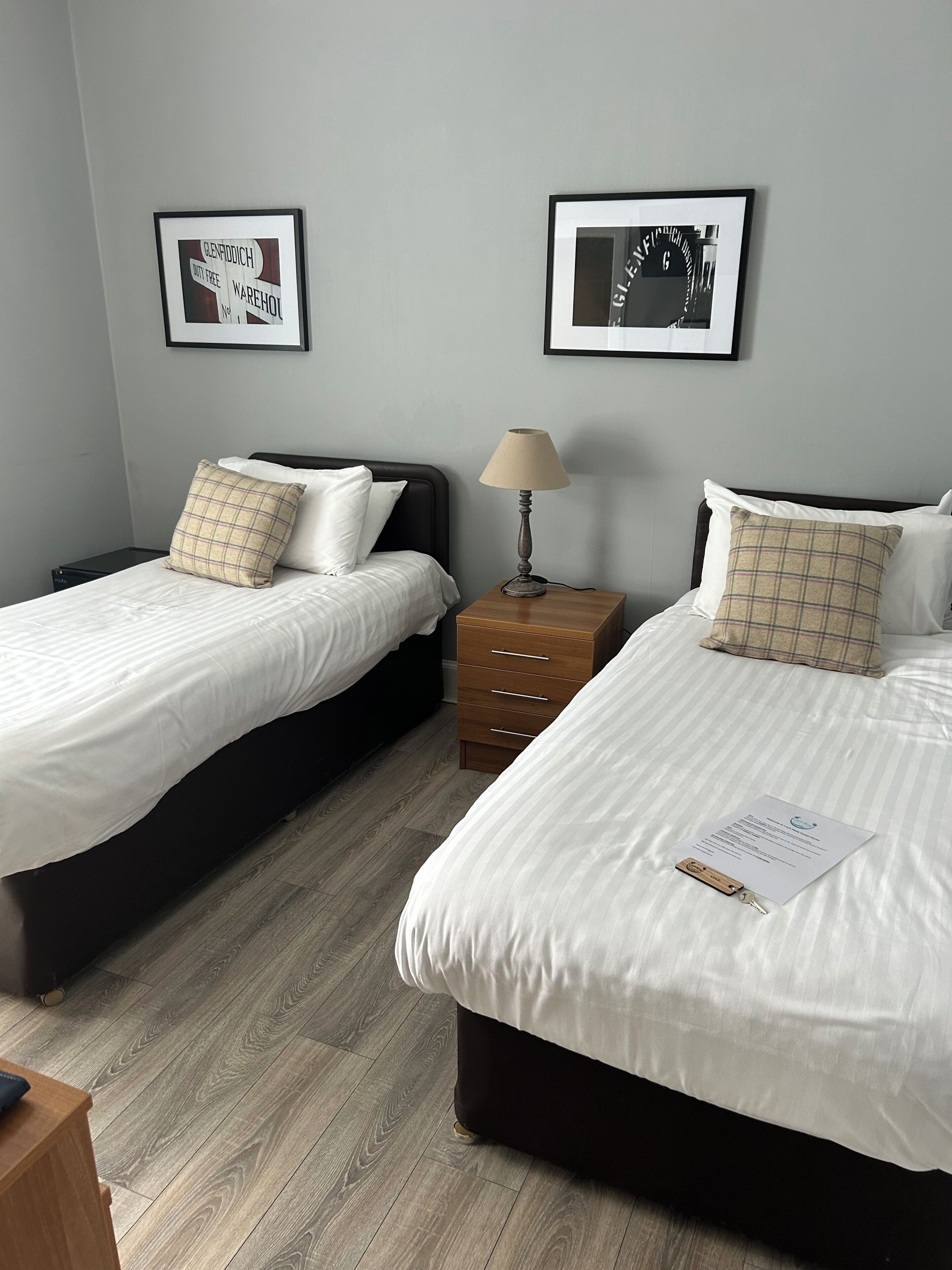 Une chambre d’hôtel avec deux lits, une table de chevet, une lampe et deux tableaux encadrés au mur.
